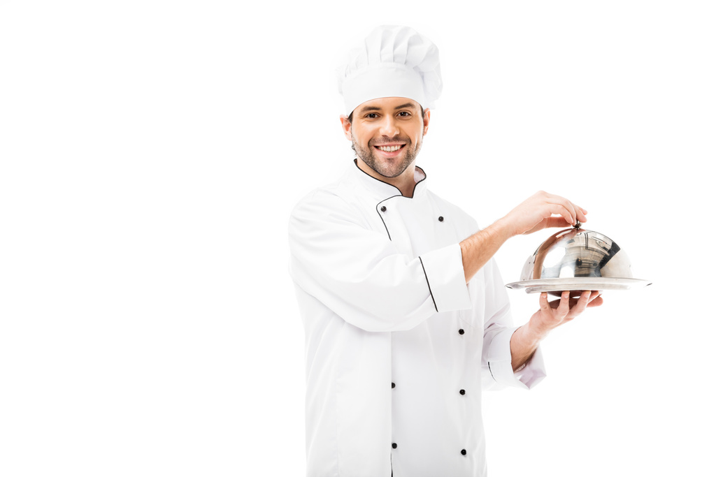 knappe jonge chef-kok holding schaal met koepel en kijken naar camera geïsoleerd op wit - Foto, afbeelding