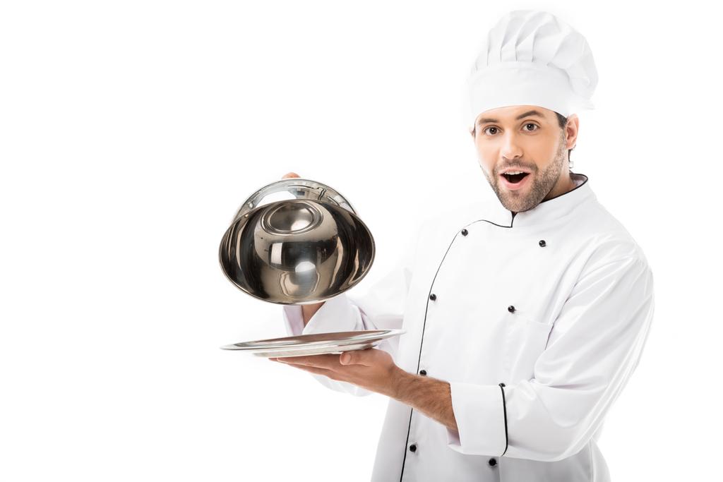 aufgeregter junger Koch, der die Kuppel vom Teller nimmt und isoliert auf weiß in die Kamera blickt - Foto, Bild