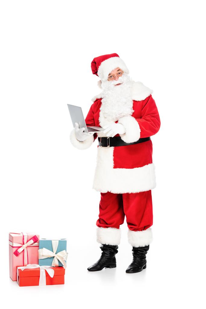 Άγιος Βασίλης με δώρα κρατώντας το laptop και βλέπουν τα φωτογραφικών μηχανών που απομονώνονται σε λευκό - Φωτογραφία, εικόνα