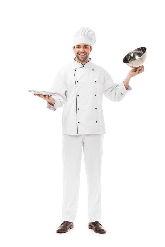 lachende jonge chef-kok nemen voor portie dome van plaat en kijken naar camera geïsoleerd op wit - Foto, afbeelding