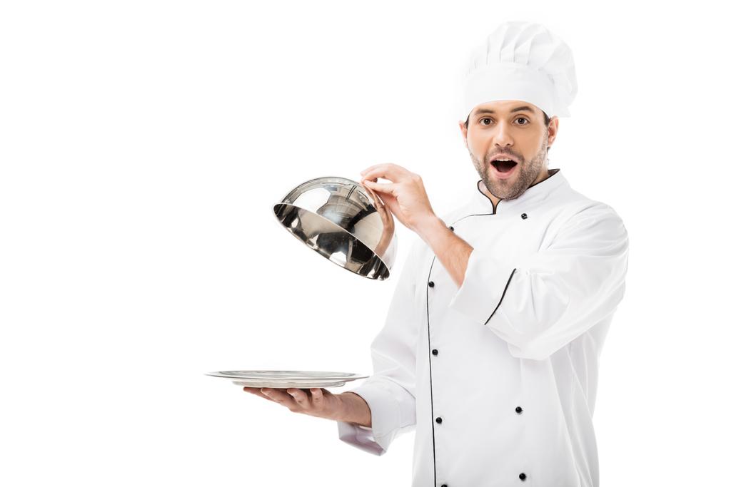überraschter junger Koch, der die Kuppel vom Teller nimmt und isoliert auf weiß in die Kamera schaut - Foto, Bild