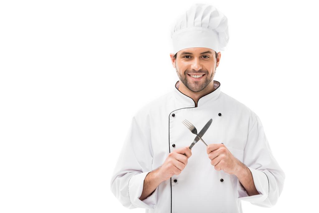 Lächelnder junger Koch, der Gabel und Messer in der Hand hält und isoliert auf weiß in die Kamera blickt - Foto, Bild