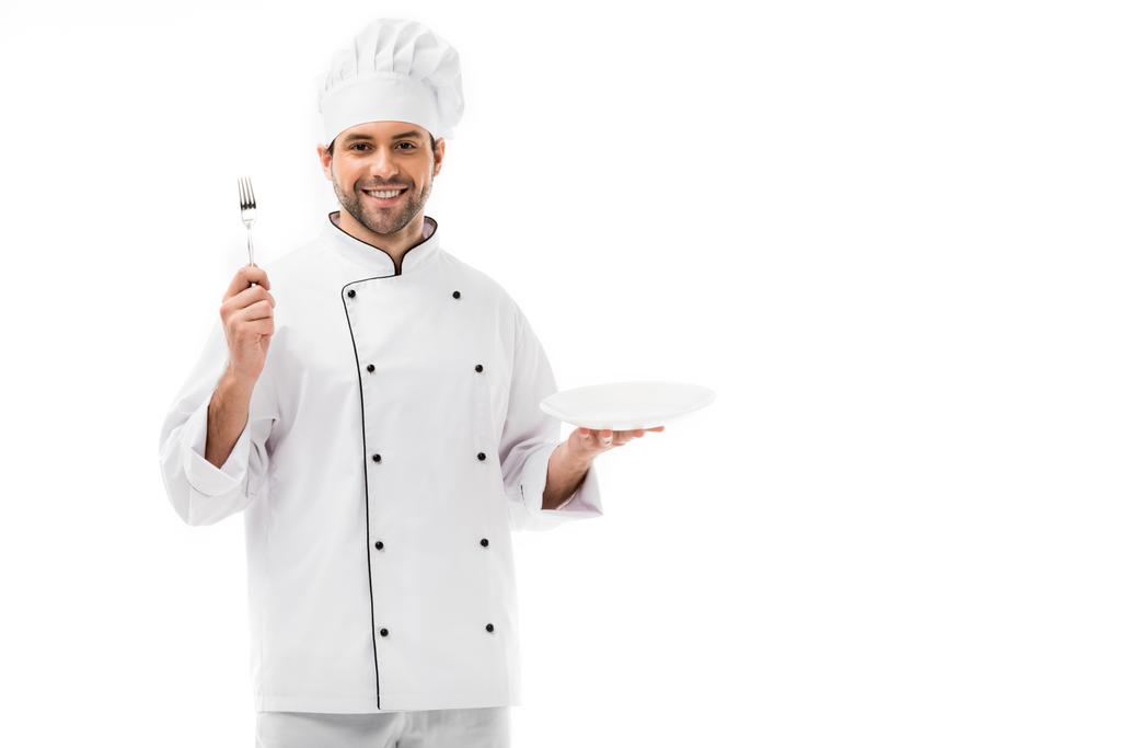 usmívající se mladý kuchař s ploténkou a vidlice při pohledu na fotoaparát izolované na bílém - Fotografie, Obrázek