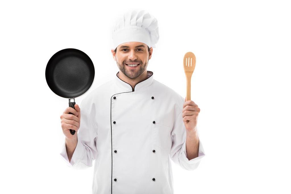 happy young chef holding frying pan and wooden spatula isolated on white - Photo, Image