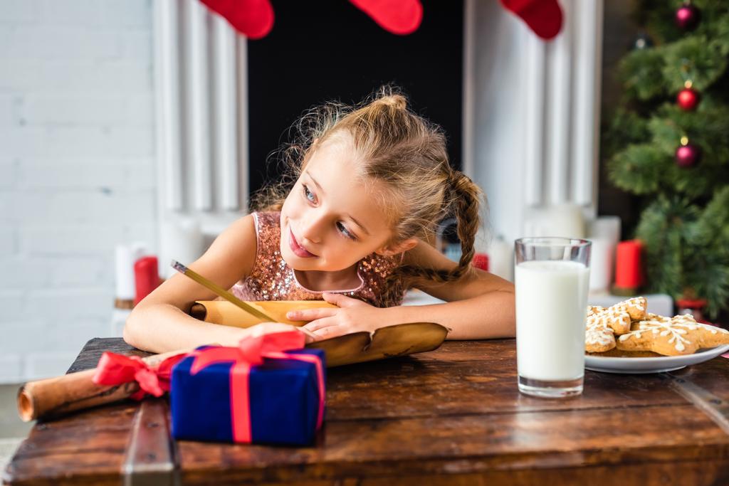 Niedliches lächelndes Kind, das wegschaut, während es zu Weihnachten Wunschliste schreibt - Foto, Bild