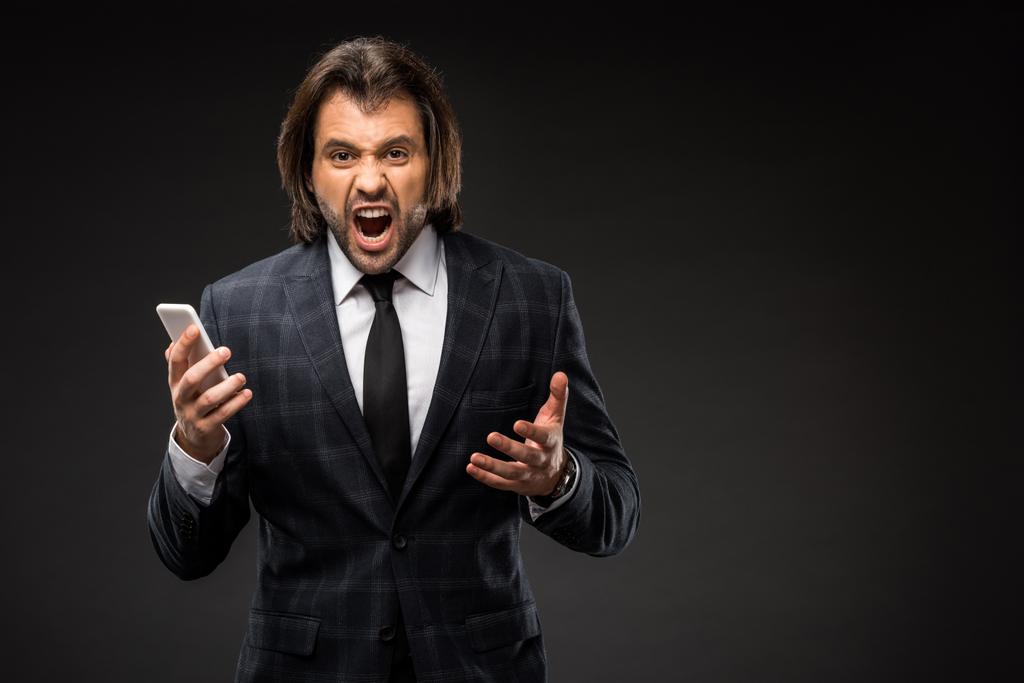 angry young businessman holding smartphone and yelling at camera isolated on black - Photo, Image
