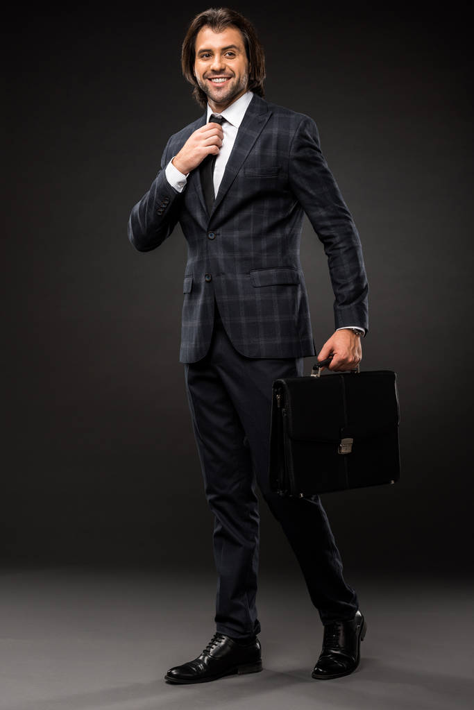 full length view of handsome young businessman holding briefcase and adjusting necktie on black - Photo, Image