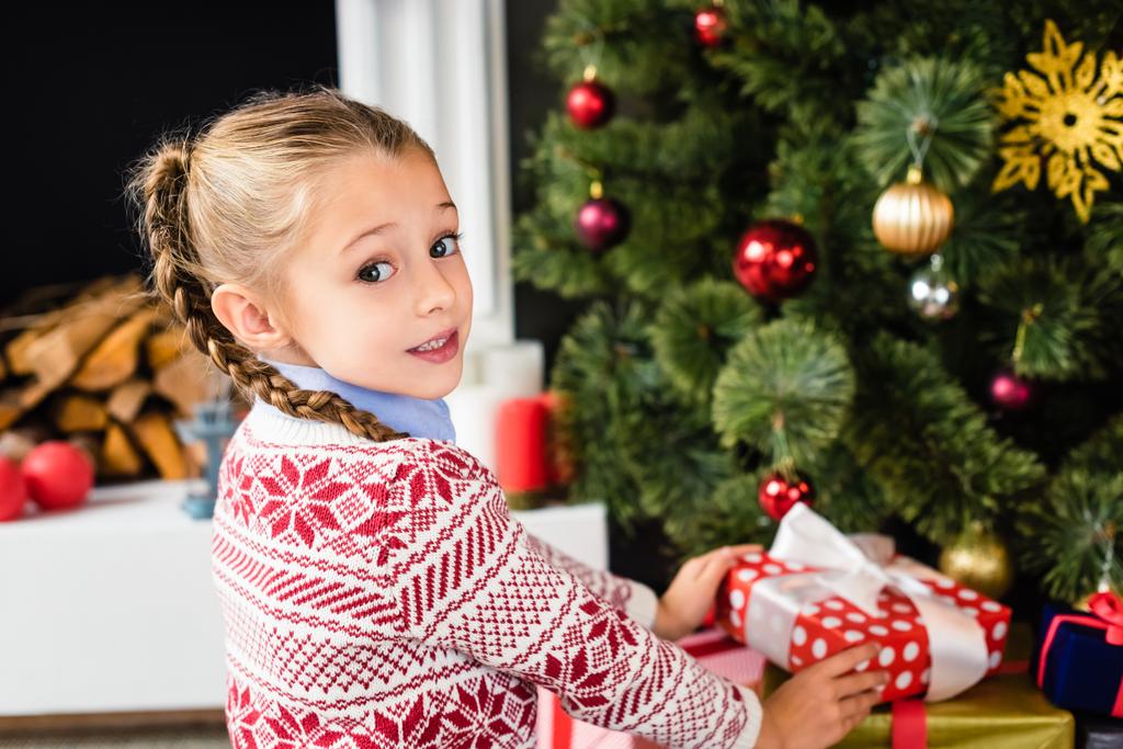 entzückendes Kind blickt in die Kamera, während es Geschenke unter dem Weihnachtsbaum hält - Foto, Bild