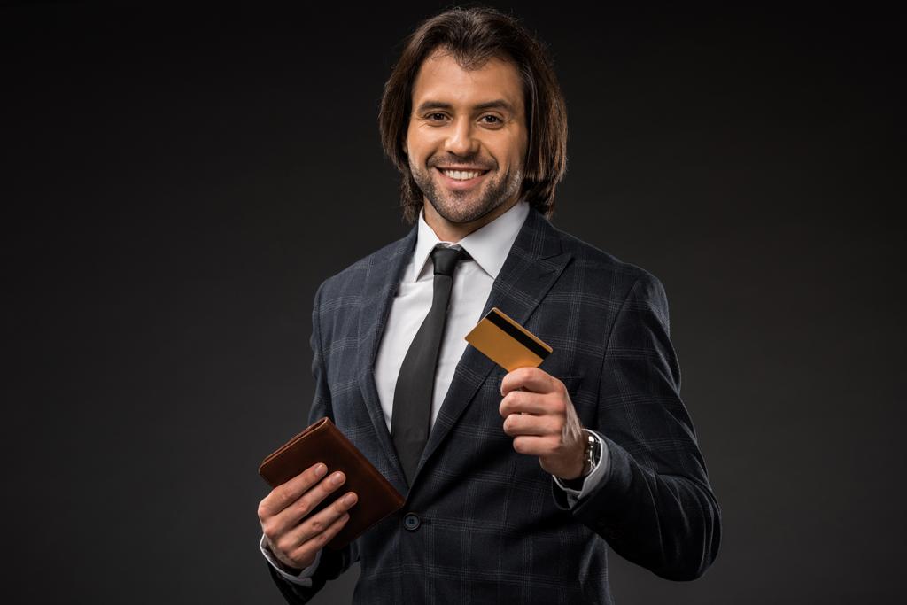 sonriente joven empresario sosteniendo cartera y tarjeta de crédito aislado en negro
  - Foto, Imagen