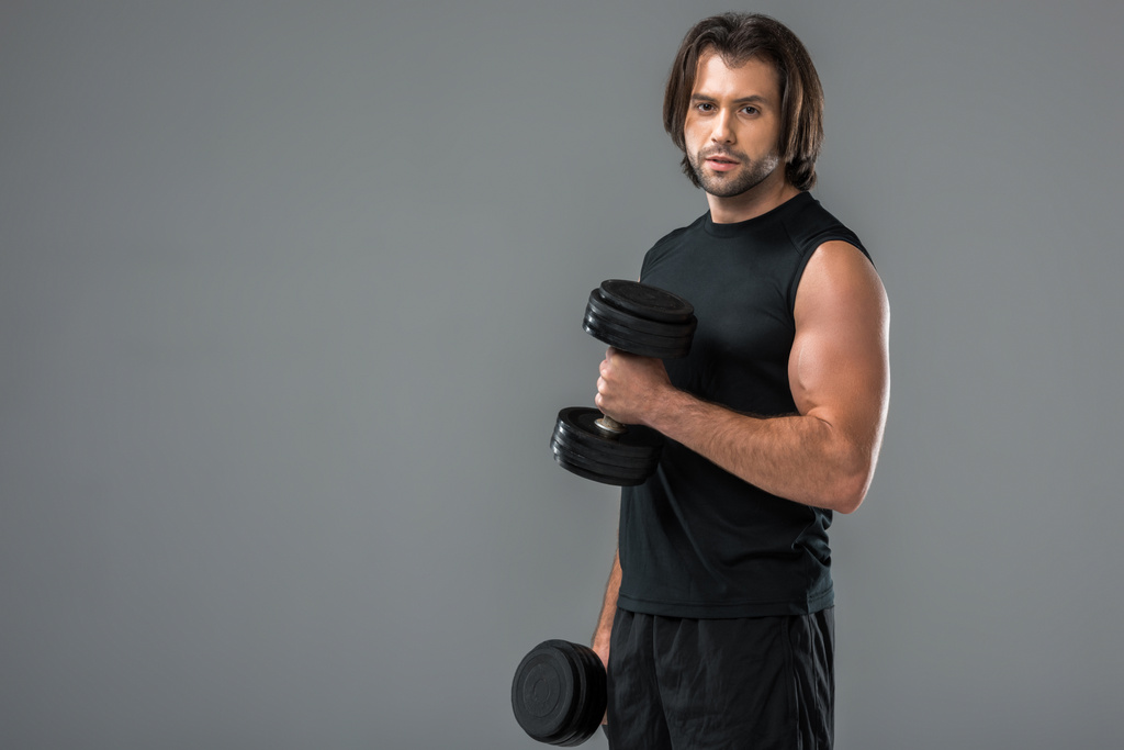 muscular young man exercising with dumbbells and looking at camera isolated on grey  - Photo, Image