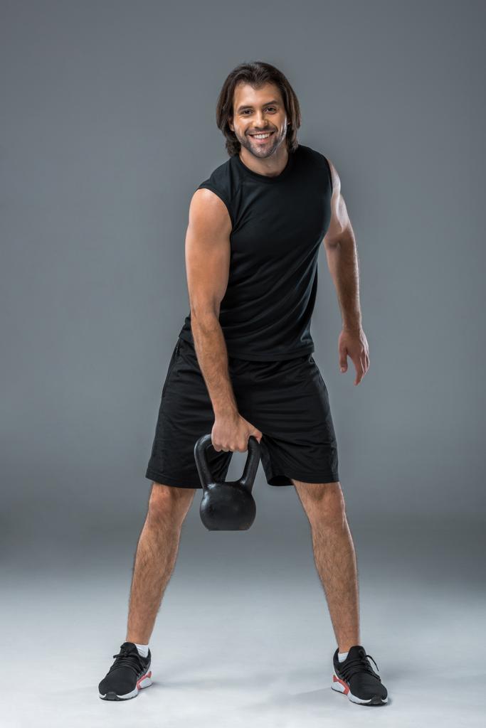 apuesto deportista sosteniendo kettlebell y sonriendo a la cámara en gris
 - Foto, imagen