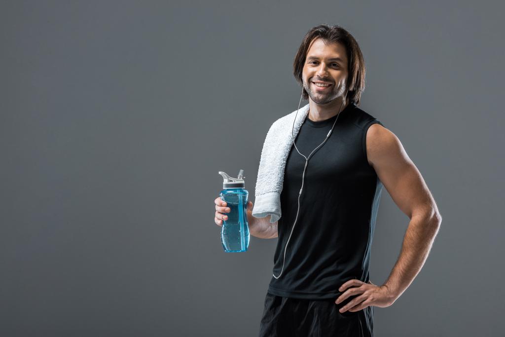 handsome young sportsman in earphones holding bottle of water and smiling at camera isolated on grey  - Photo, Image