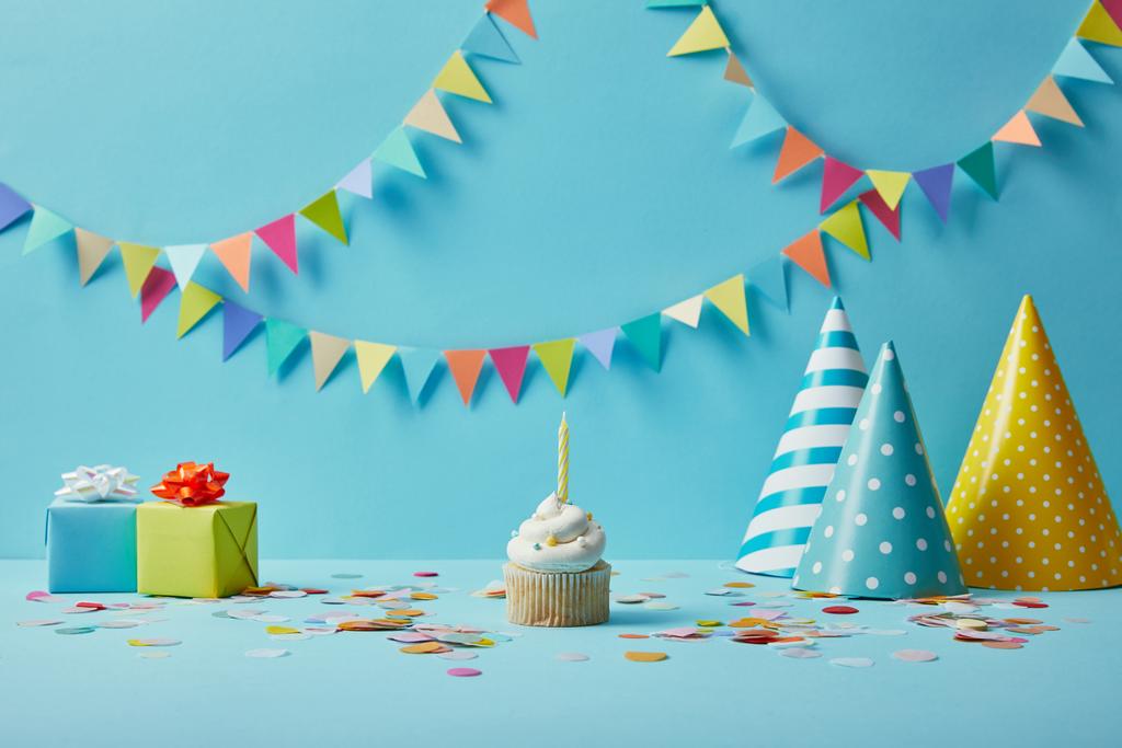  Delicious cupcake, party hats, confetti and gifts on blue background with colorful bunting - Photo, Image