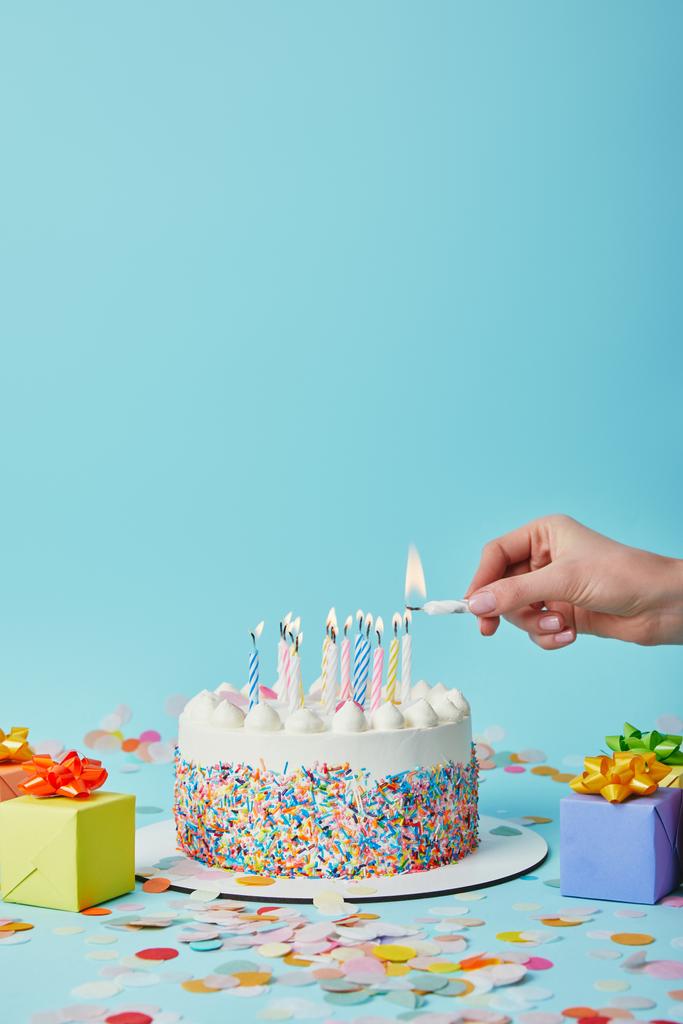 Partial view of woman lighting birthday cake on blue background with gifts and confetti - Photo, Image
