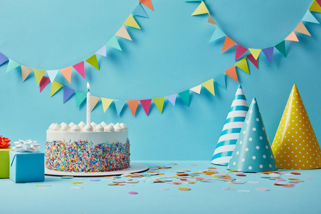 Heerlijke verjaardagstaart, geschenken, feestmutsen en confetti op blauwe achtergrond met bunting - Foto, afbeelding