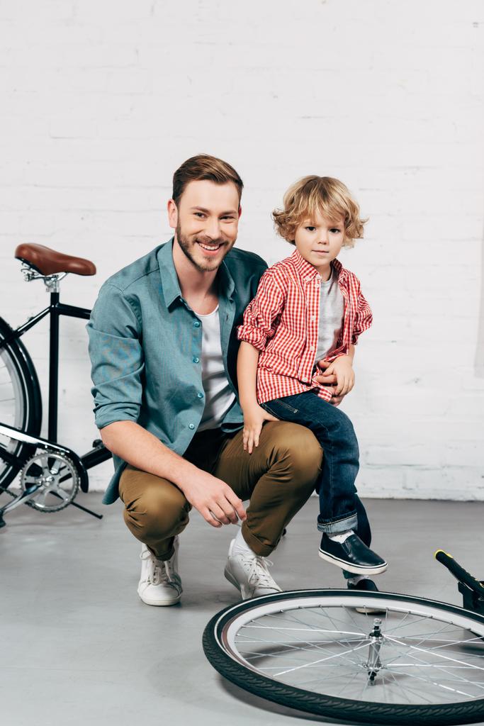 ευτυχισμένος άνθρωπος κάθεται με το μικρό του γιο και κοιτάζοντας κάμερα σε συνεργείο ποδηλάτων - Φωτογραφία, εικόνα