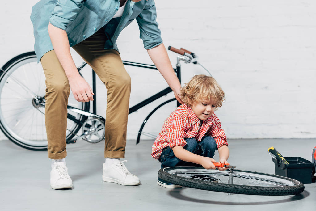 körülvágott kép az ember közelében kis fia által fogó kerék rögzítő kerékpár műhely - Fotó, kép