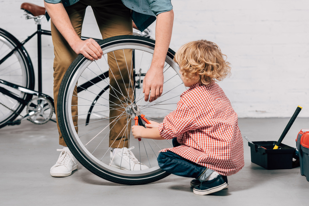cropped image of father holding bicycle wheel while his little son repairing with pliers at workshop - Photo, Image