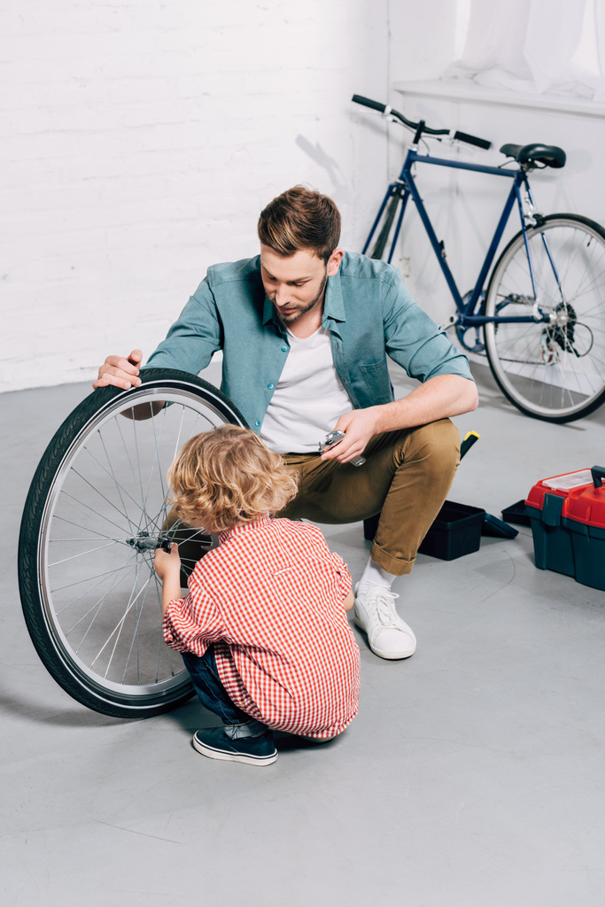 vista de ángulo alto del padre sosteniendo la rueda de bicicleta mientras su pequeño hijo reparando en el taller
 - Foto, Imagen