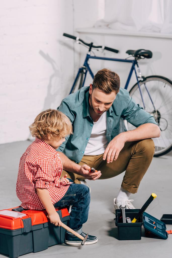 man holding pliers and sitting with little son sitting near opened tools boxes in bicycle workshop - Photo, Image