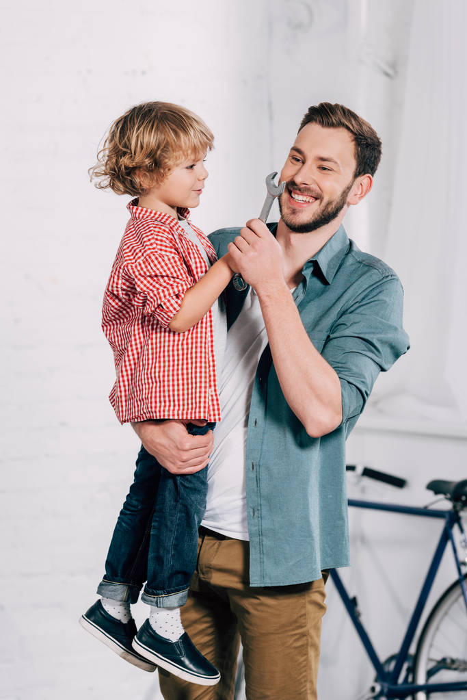 Lachender Mann hält Sohn mit Schraubenschlüssel in der Hand in Werkstatt  - Foto, Bild
