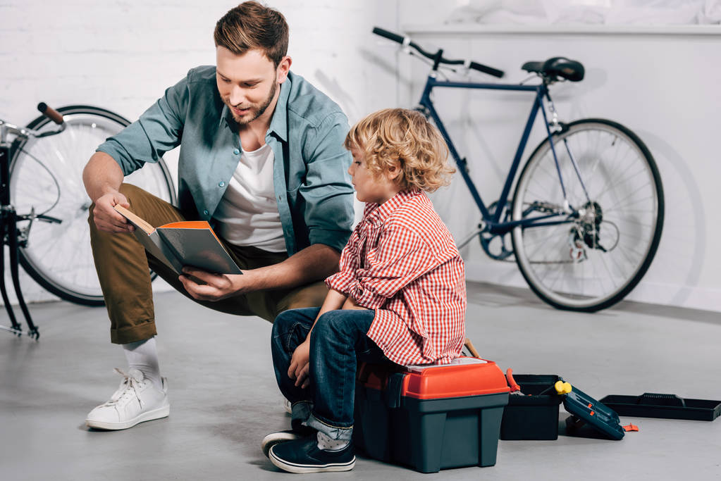 Χαμογελαστός άνθρωπος ανάγνωση οδηγίες στο μικρό γιο κάθεται σε εργαλεία πλαίσιο στο συνεργείο ποδηλάτων - Φωτογραφία, εικόνα