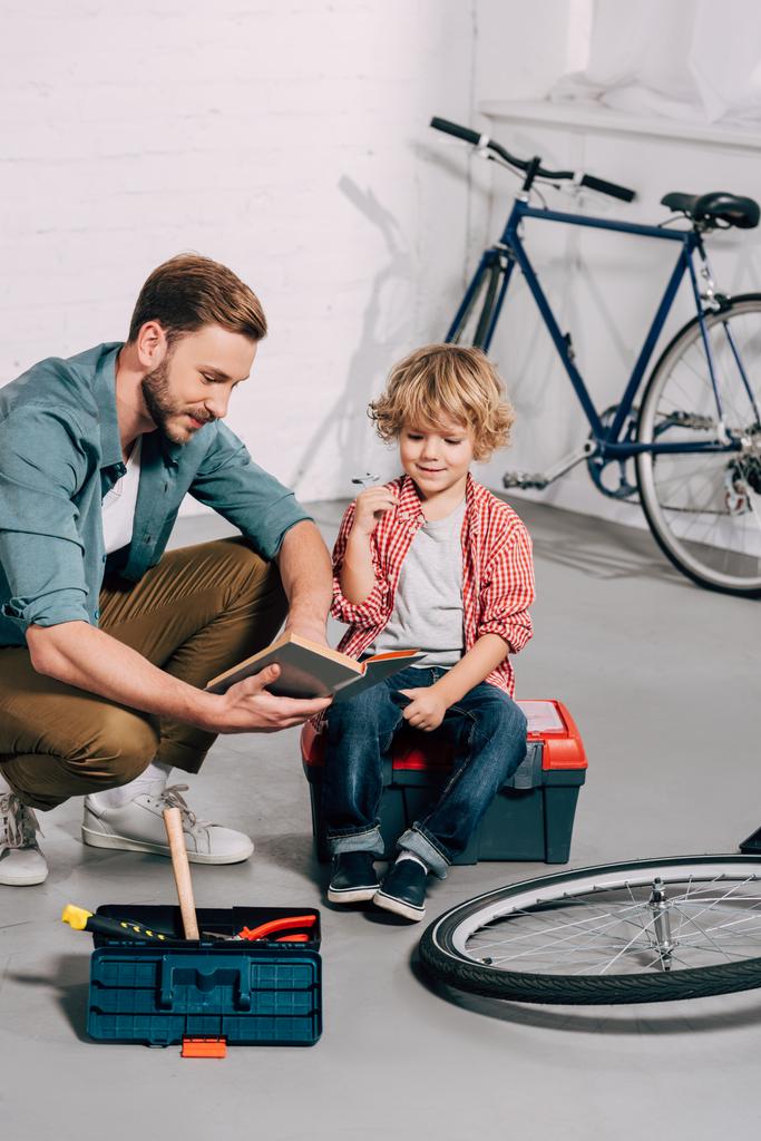 happy man pointing by hand at book to little son sitting on tools box in bicycle workshop  - Photo, Image
