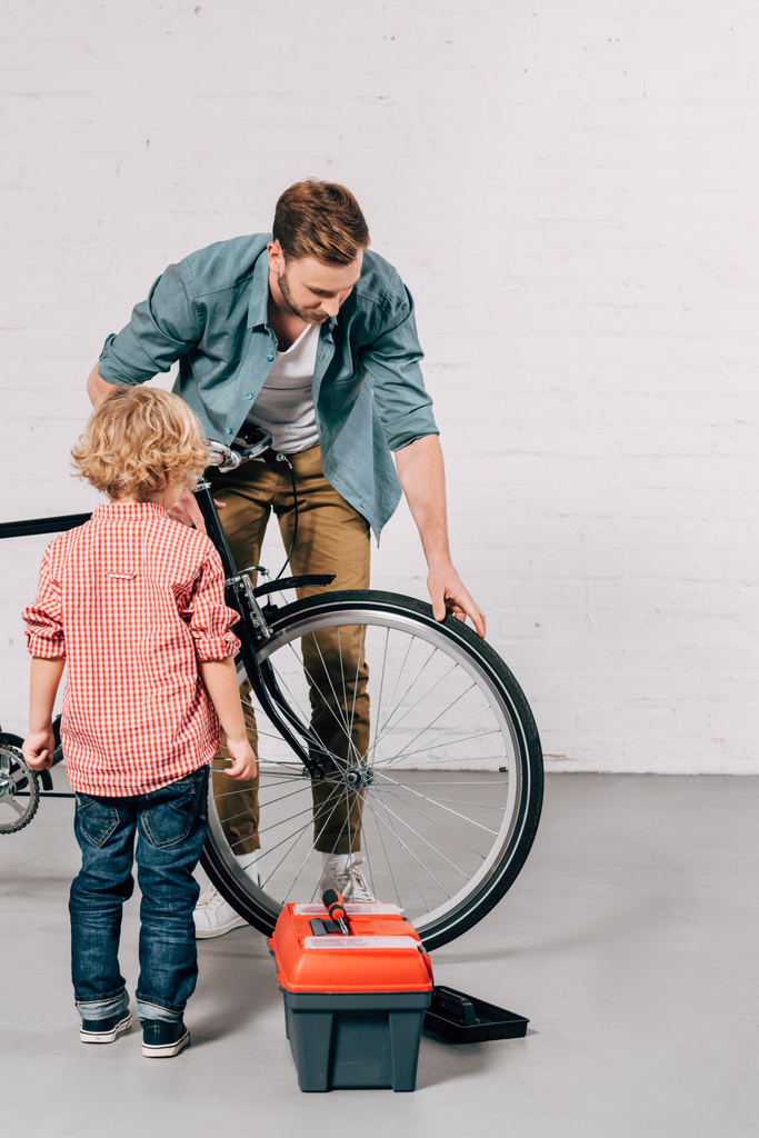 Отец показывает маленькому сыну, как починить велосипед возле ящика с инструментами в мастерской
 - Фото, изображение