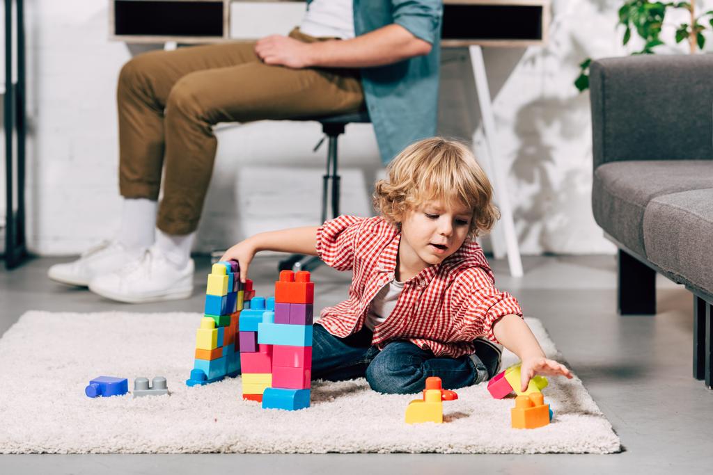 petit garçon bouclé jouant avec des blocs de plastique sur le sol tandis que son père assis derrière à la maison
 - Photo, image