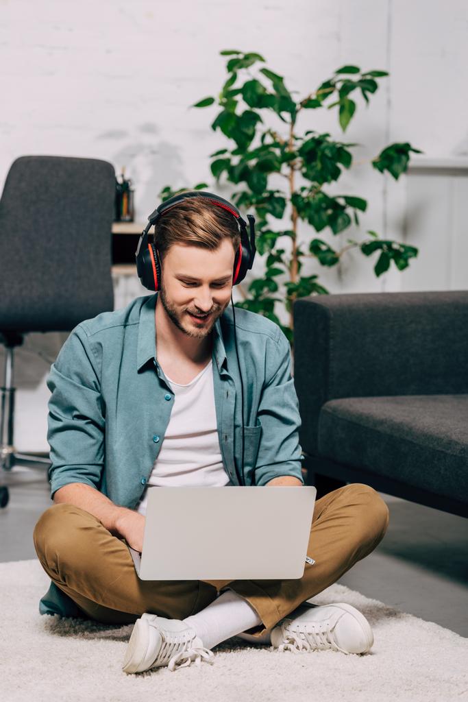sonriente freelancer masculino en auriculares que trabajan en el ordenador portátil mientras está sentado en el suelo en casa
 - Foto, imagen