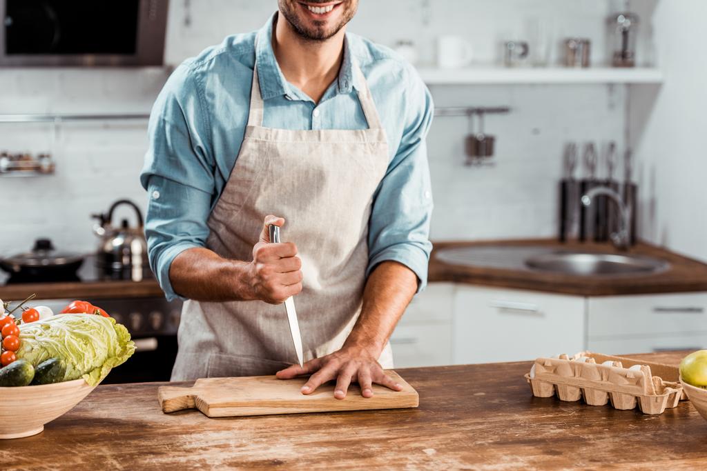 Schnappschuss eines lächelnden jungen Mannes in Schürze mit Messer und Hand auf Schneidebrett in Küche - Foto, Bild