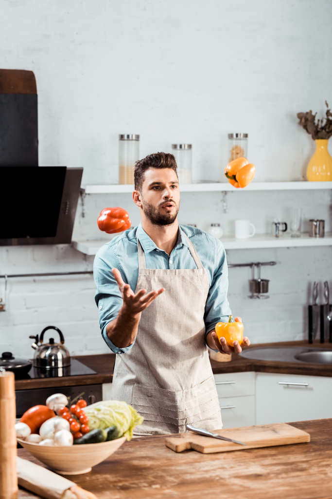 handsome young man in apron juggling with fresh peppers in kitchen - Photo, Image