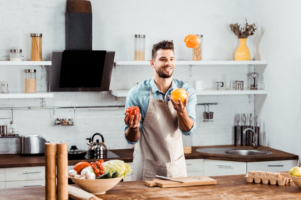 guapo joven sonriente en delantal sosteniendo verduras frescas en la cocina
 - Foto, imagen