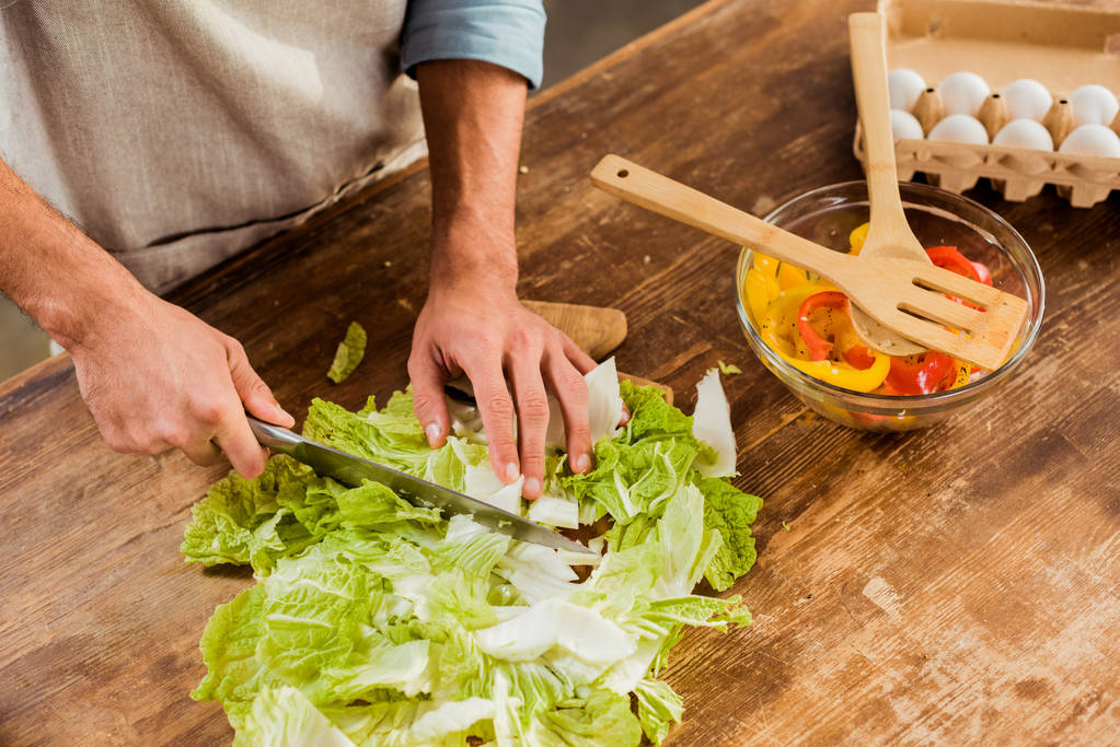 обрезанный снимок человека в фартуке, рубящей капусту и готовящем овощной салат
 - Фото, изображение