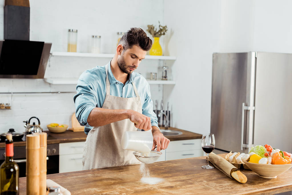 schöner junger Mann in Schürze sichtet Mehl auf Küchentisch  - Foto, Bild