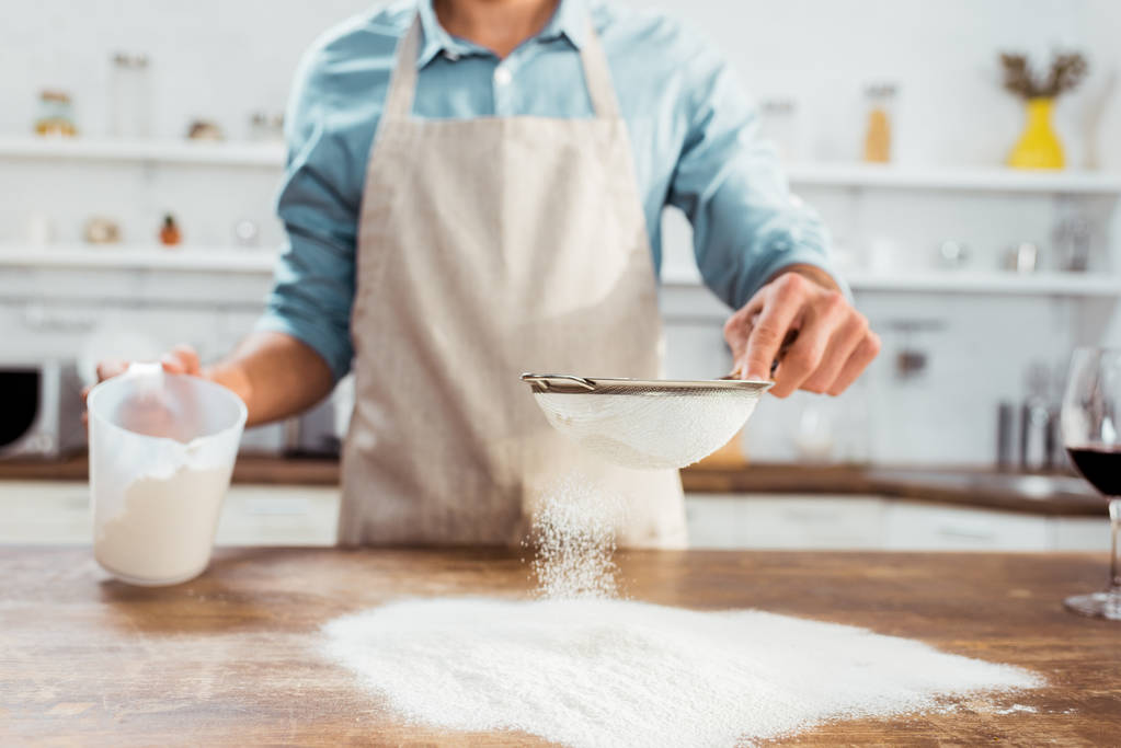 częściowy widok młodego człowieka w fartuch przesiewania mąki na stole w kuchni  - Zdjęcie, obraz