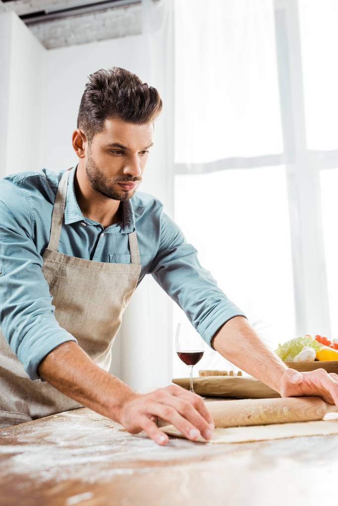 エプロン麺棒を押しながら台所で生地の準備でハンサムな若い男 - 写真・画像