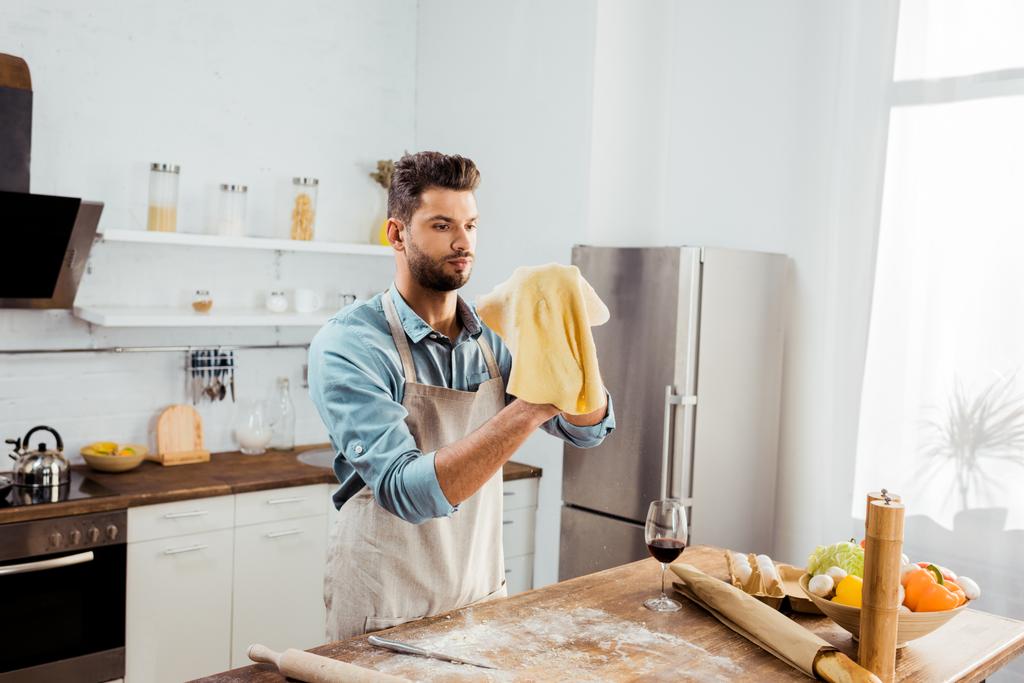 joven guapo en delantal sosteniendo masa cruda mientras prepara pizza
  - Foto, Imagen