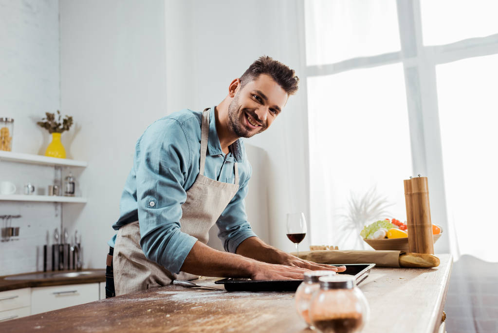 csinos, fiatal férfi-kötény mosolyogva kamera felkészülése pizza tésztát sütés tálca - Fotó, kép