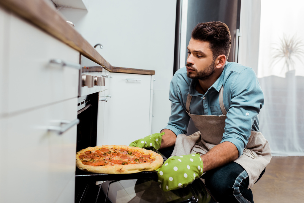 Молодой человек в фартуке и с выбоинами достает поднос для выпечки пиццей из духовки
 - Фото, изображение