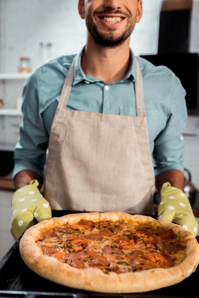 plan recadré d'un homme souriant dans un tablier et des poignées tenant une plaque de cuisson avec une pizza fraîche maison
 - Photo, image