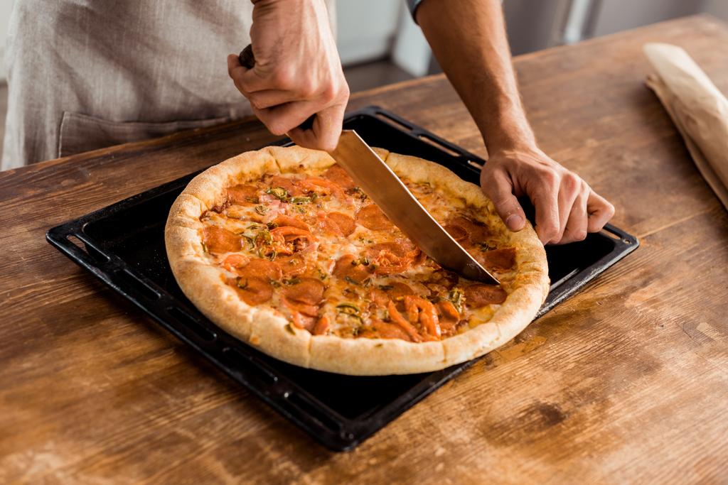 vista parcial del hombre con cuchillo cortando pizza fresca casera en bandeja para hornear
 - Foto, Imagen