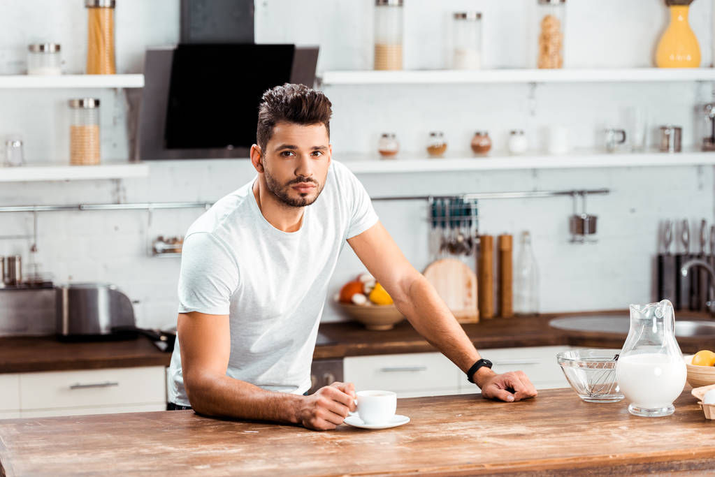 νεαρός άνδρας κρατώντας φλιτζάνι καφέ και να βλέπουν το πρωί από την φωτογραφική μηχανή στην κουζίνα - Φωτογραφία, εικόνα
