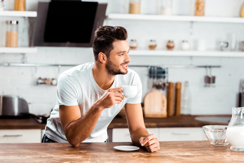 一杯のコーヒーを保持していると朝の台所のテーブルで傾いている若い男の笑みを浮かべてください。 - 写真・画像