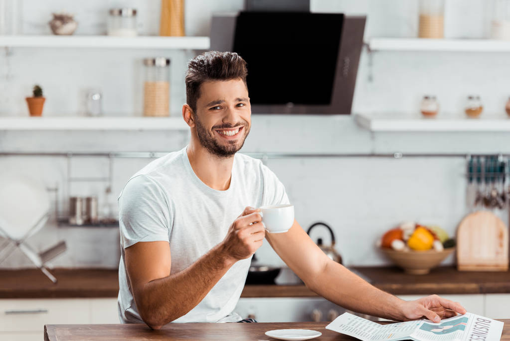 knappe jonge man houden van kopje koffie en glimlachend in de camera in de keuken - Foto, afbeelding