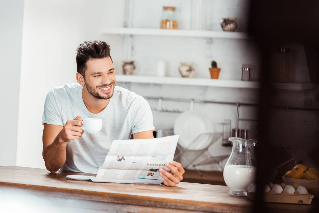 enfoque selectivo del joven sonriente sosteniendo una taza de café y leyendo el periódico en la cocina por la mañana
 - Foto, imagen