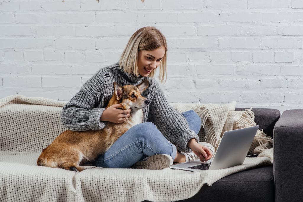 uśmiechnięta Młoda Blondynka kobieta siedzi z welsh corgi psa na kanapie i komputera przenośnego - Zdjęcie, obraz