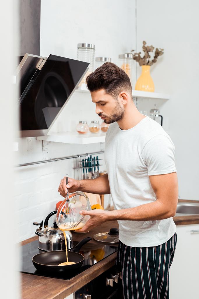 Красивый молодой человек в пижаме готовит омлет на сковородке
 - Фото, изображение