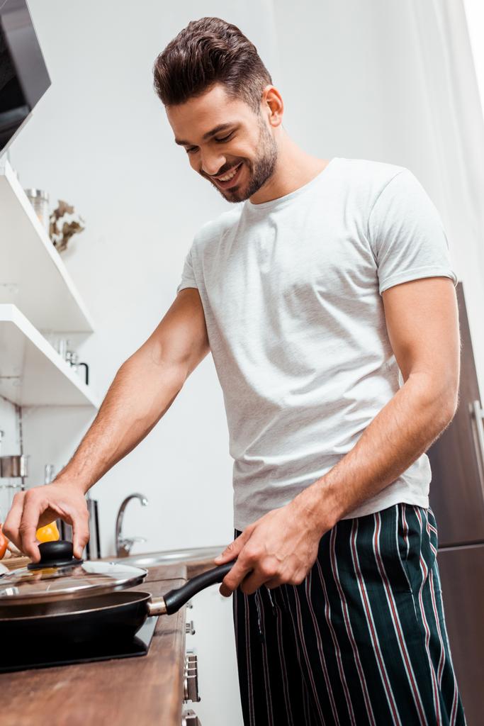 niski kąt widzenia uśmiechający się młody człowiek w piżamie omlet na patelni do gotowania - Zdjęcie, obraz