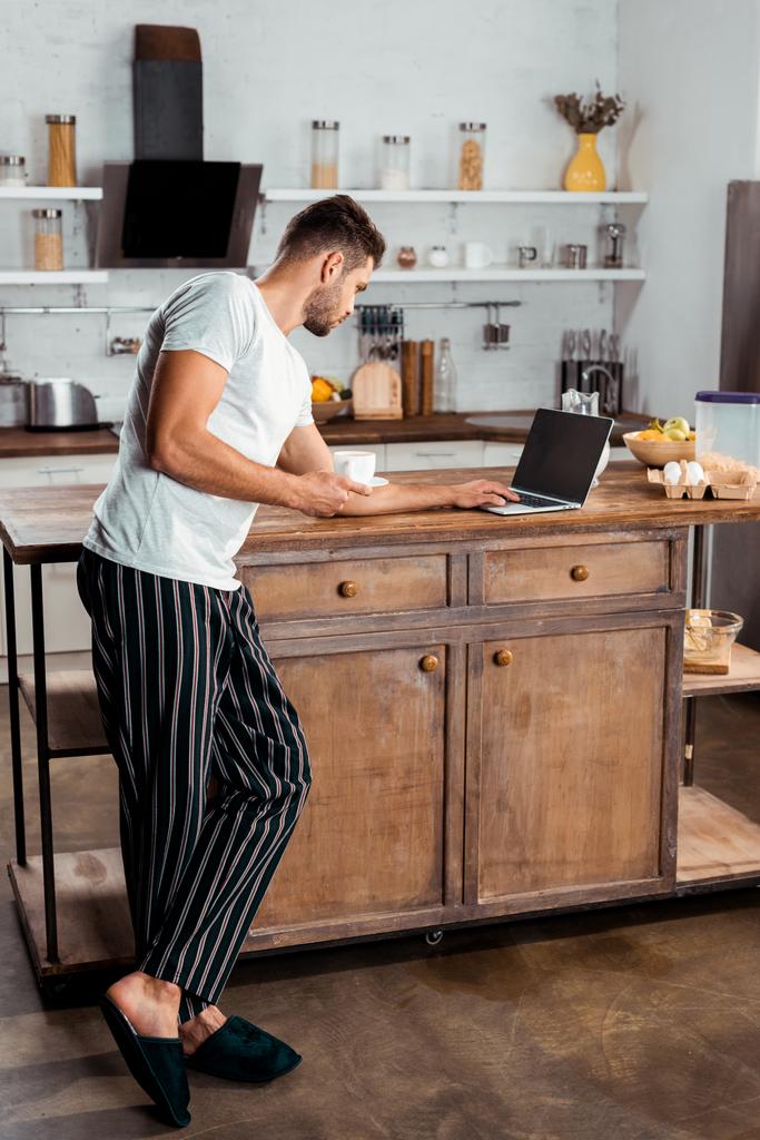 молодой человек в пижаме с помощью ноутбука и держа чашку кофе на кухне
 - Фото, изображение